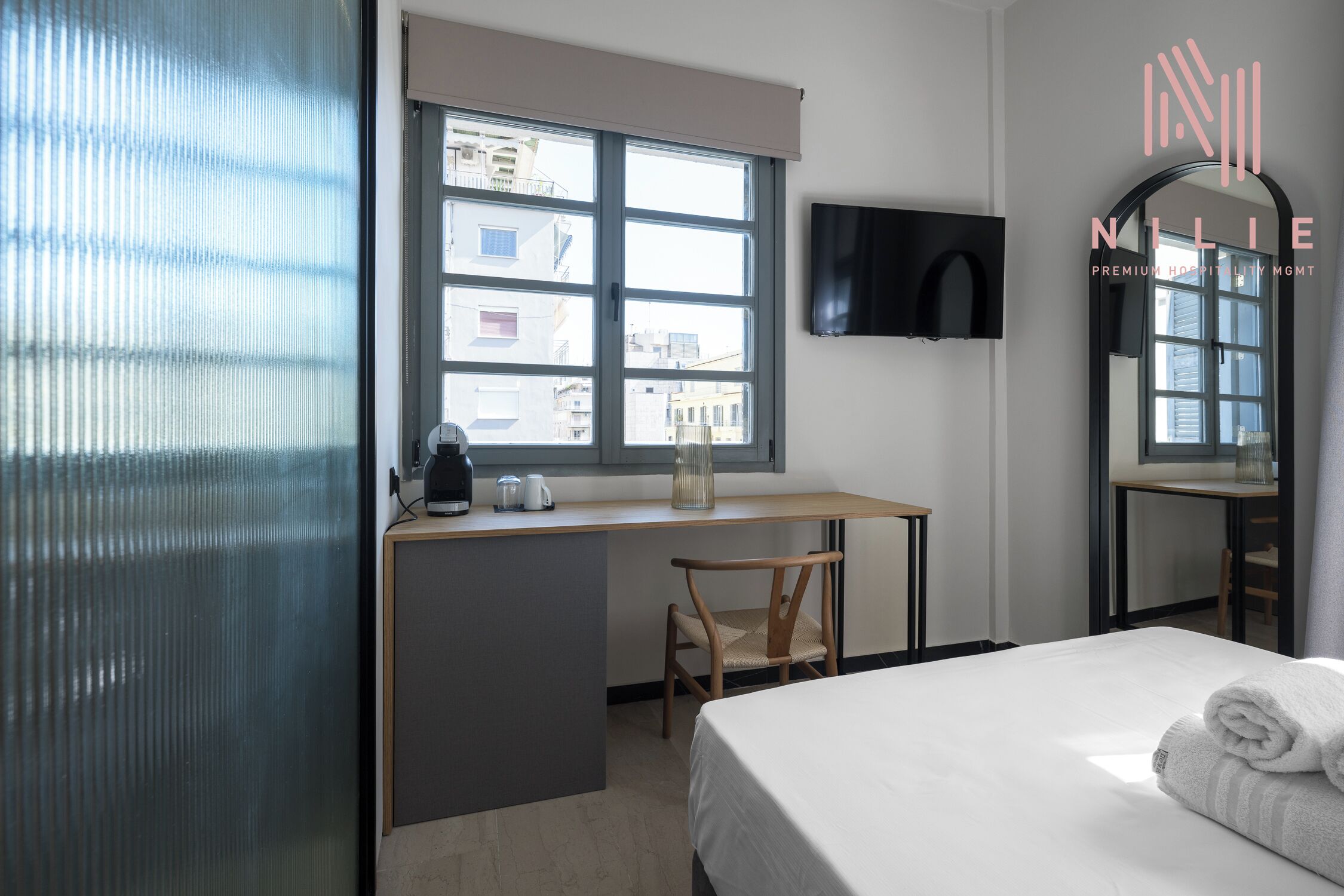 A hotel room with a bed, a desk with a chair, a TV mounted on the wall, and a window with a view of buildings.