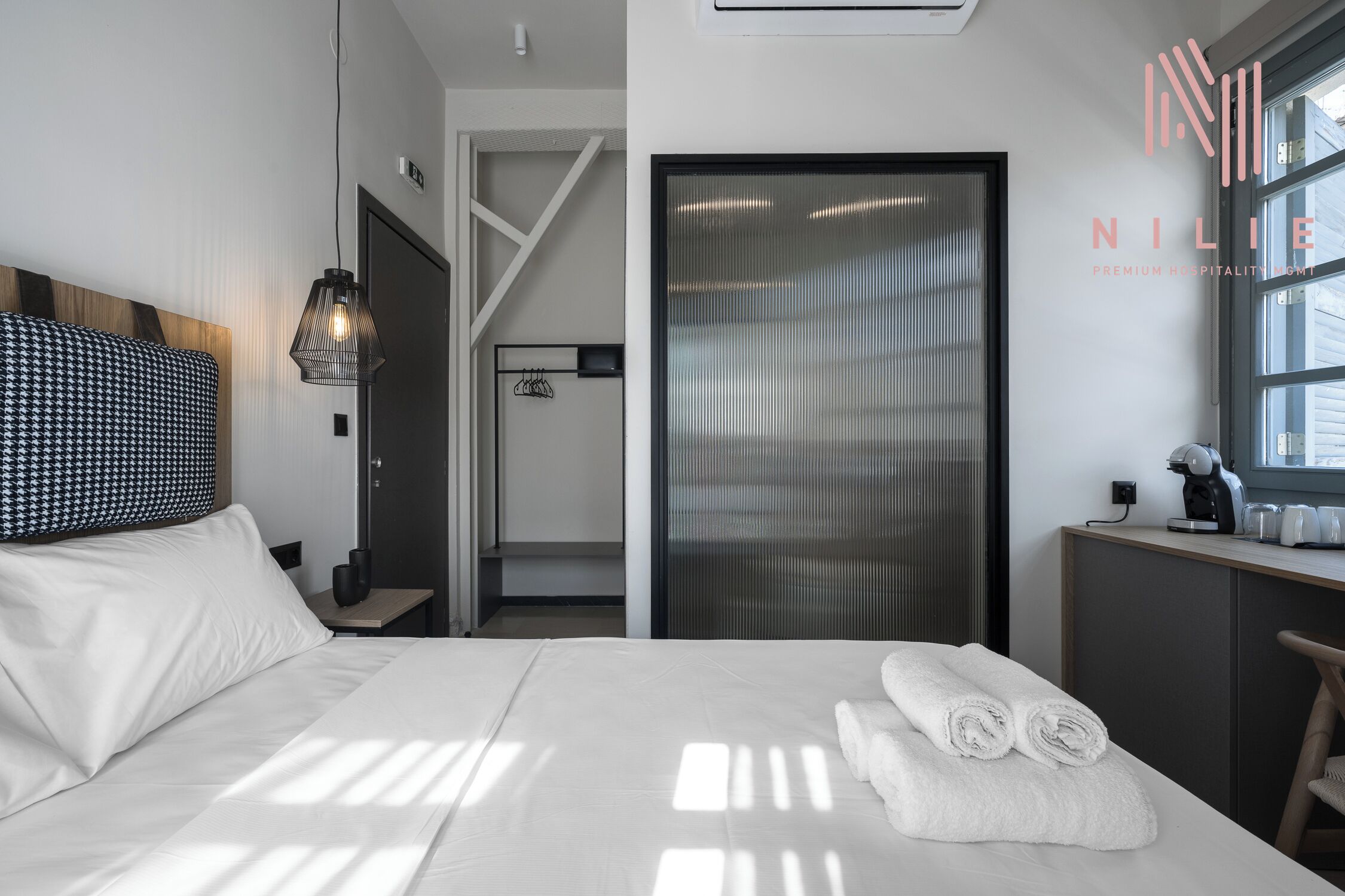 A modern hotel room with a neatly made bed, a wooden headboard, a desk with a lamp, and a view of the outside through a window.