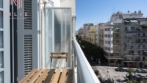 A balcony with a wooden bench, a window with blinds, and a view of a city street with buildings.