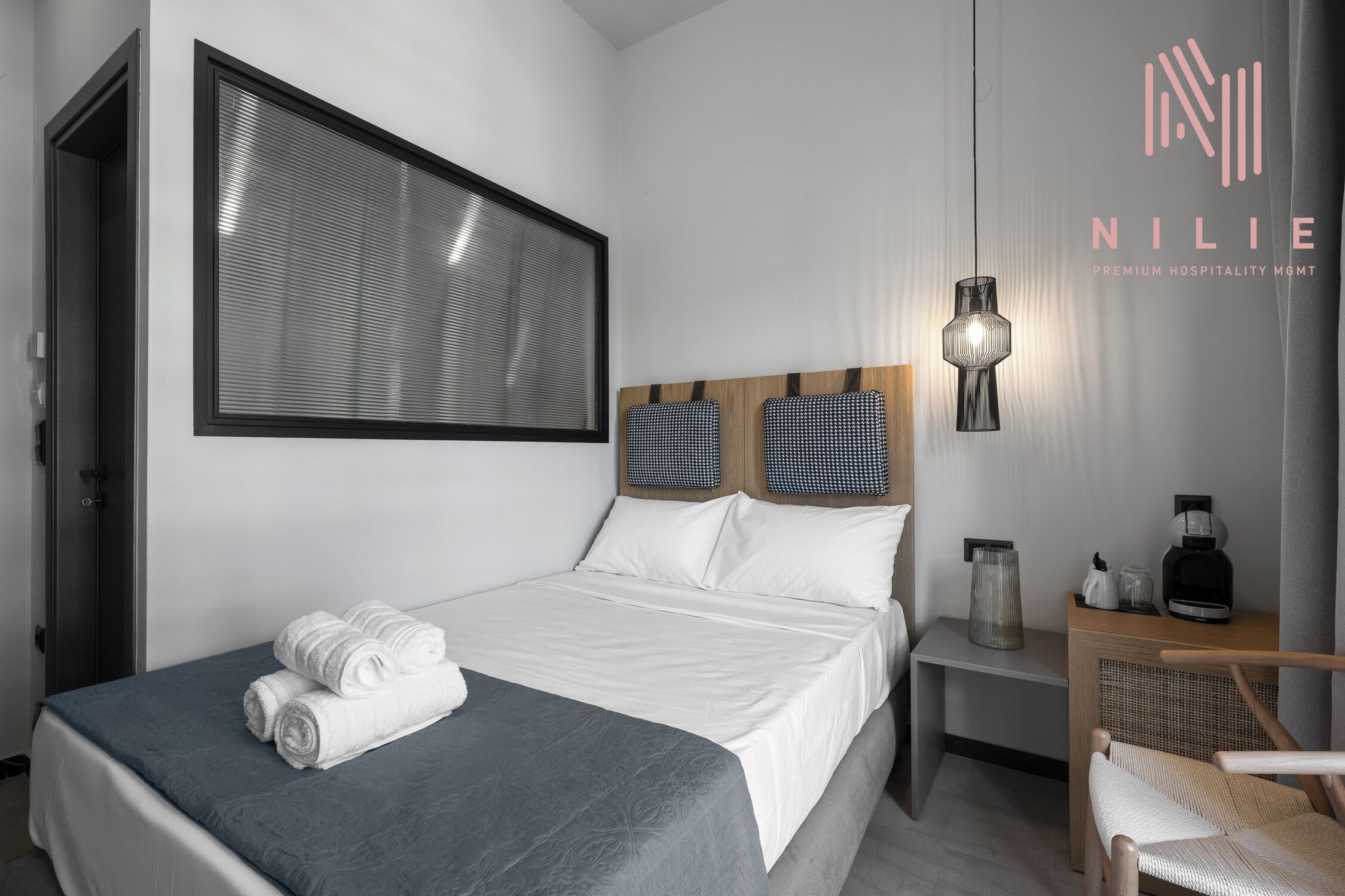 A modern hotel room with a neatly made bed, a large window with blinds, a bedside table with a lamp, and a chair.