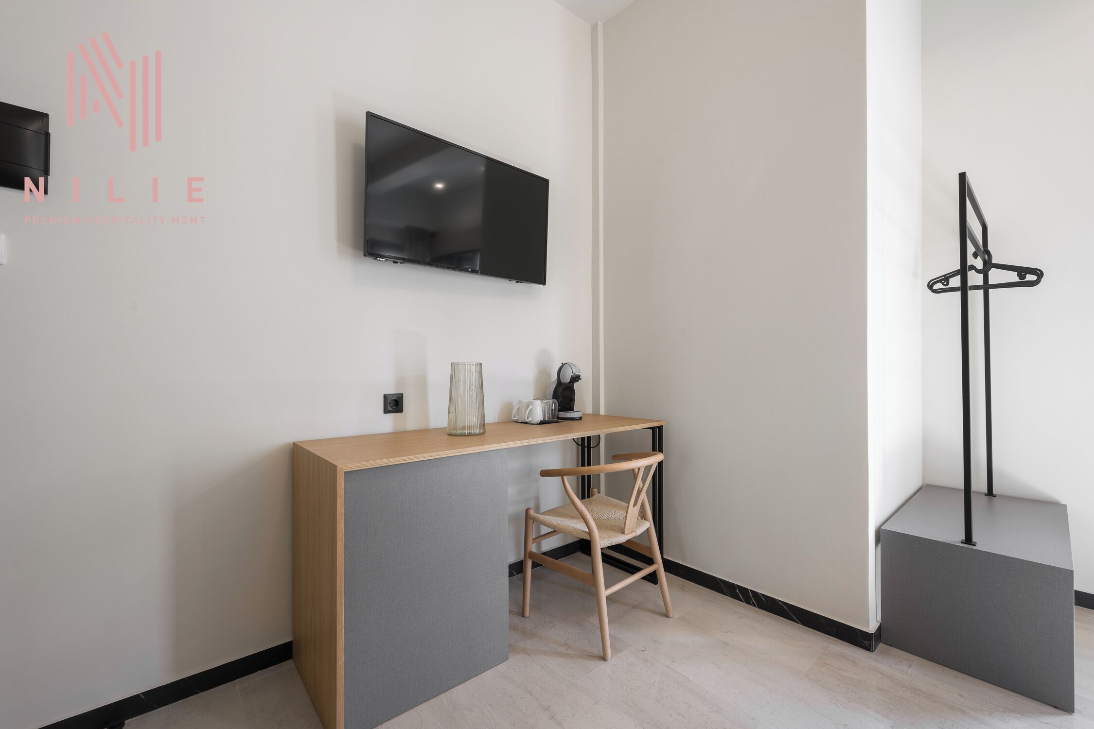 Et moderne, minimalistisk værelse med et fladskærms-tv på væggen, et træskrivebord med stol og et knagered.