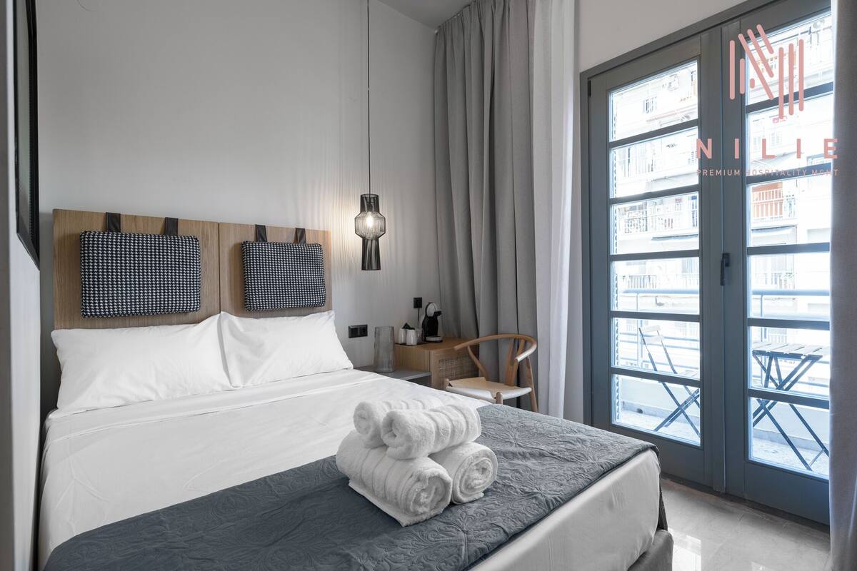 Habitación de hotel con cama, escritorio, silla, ventana con cortinas y vista a edificios.