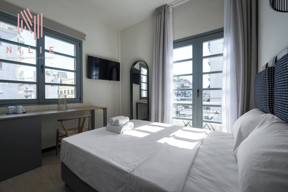 Habitación de hotel con cama, escritorio, silla, televisor y ventanas amplias con vista a la ciudad.