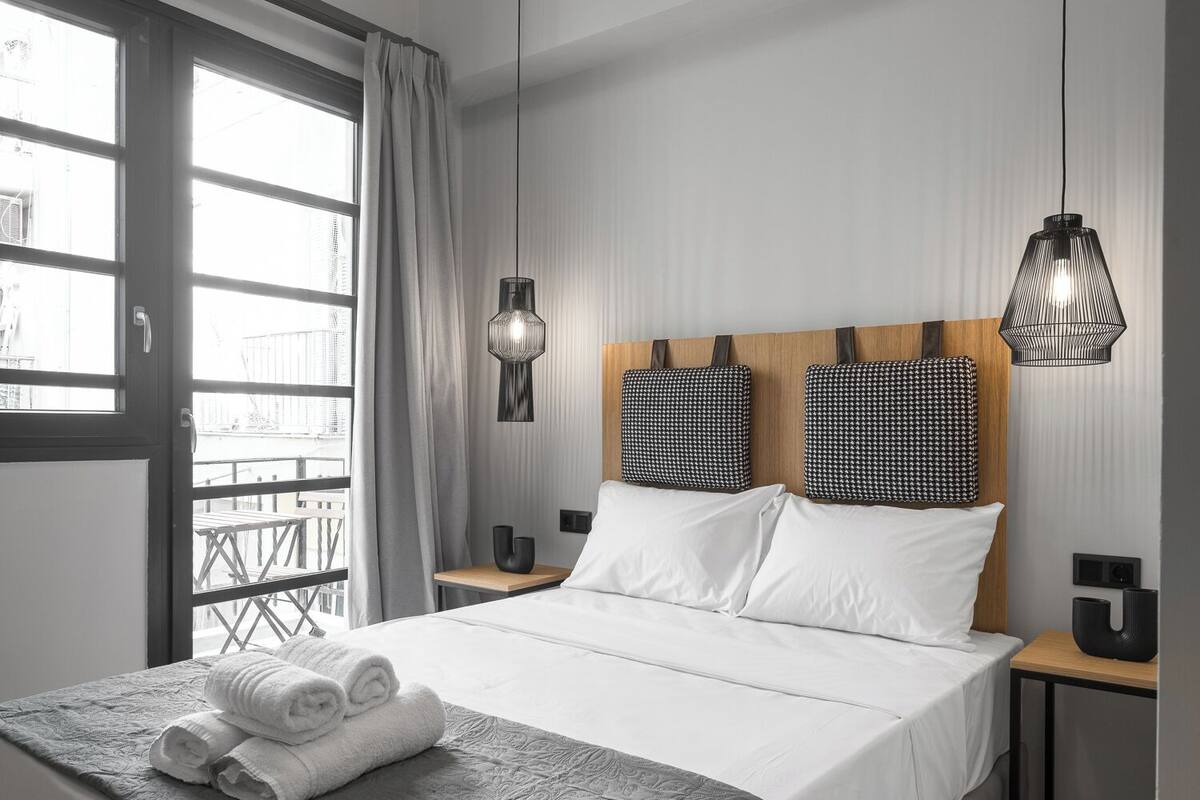 Una habitación de hotel moderna con una cama grande, dos mesitas de noche, una ventana con cortinas y vistas al balcón.