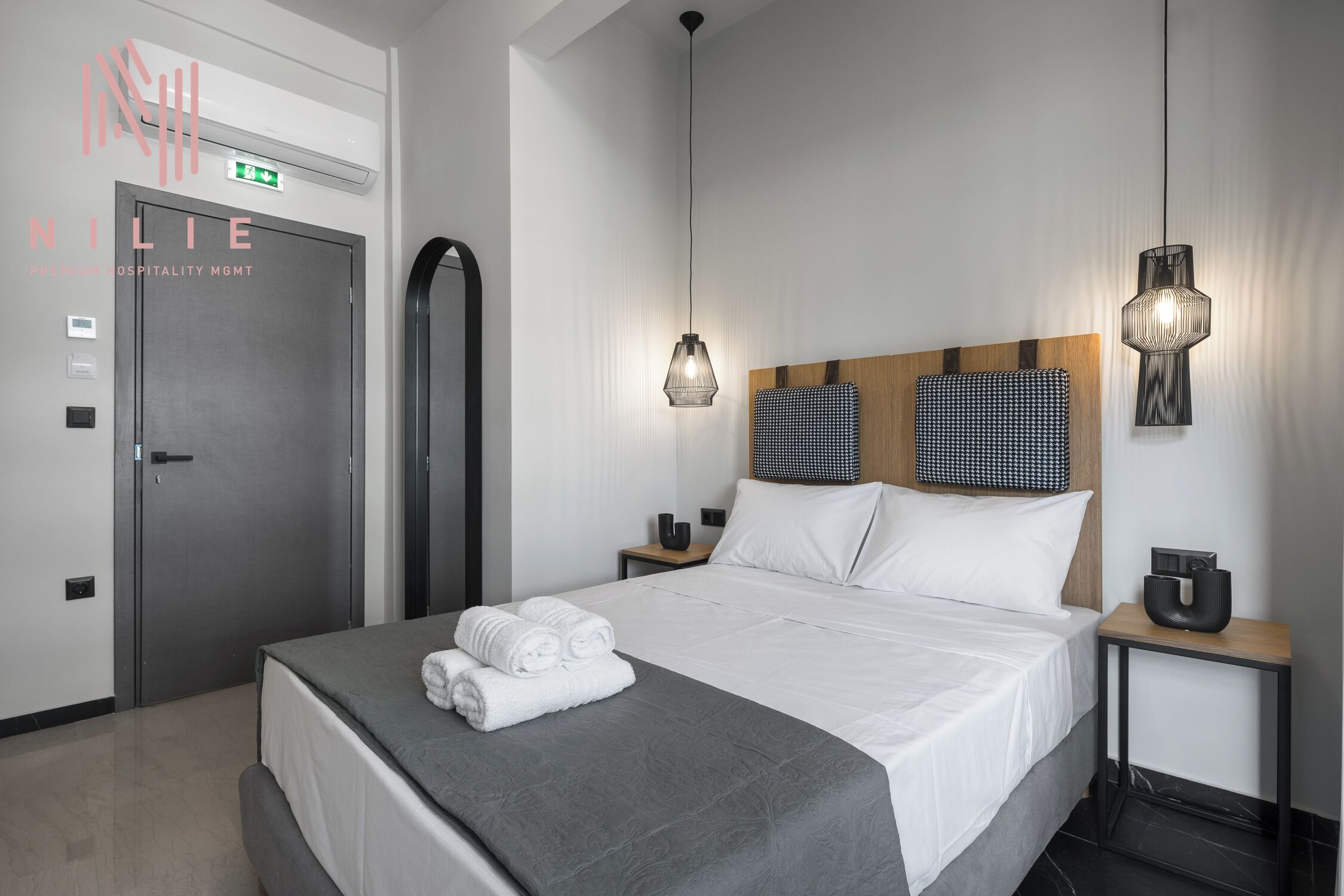 A modern hotel room with a neatly made bed, a wooden headboard, two bedside tables with lamps, and a door with an emergency exit sign.