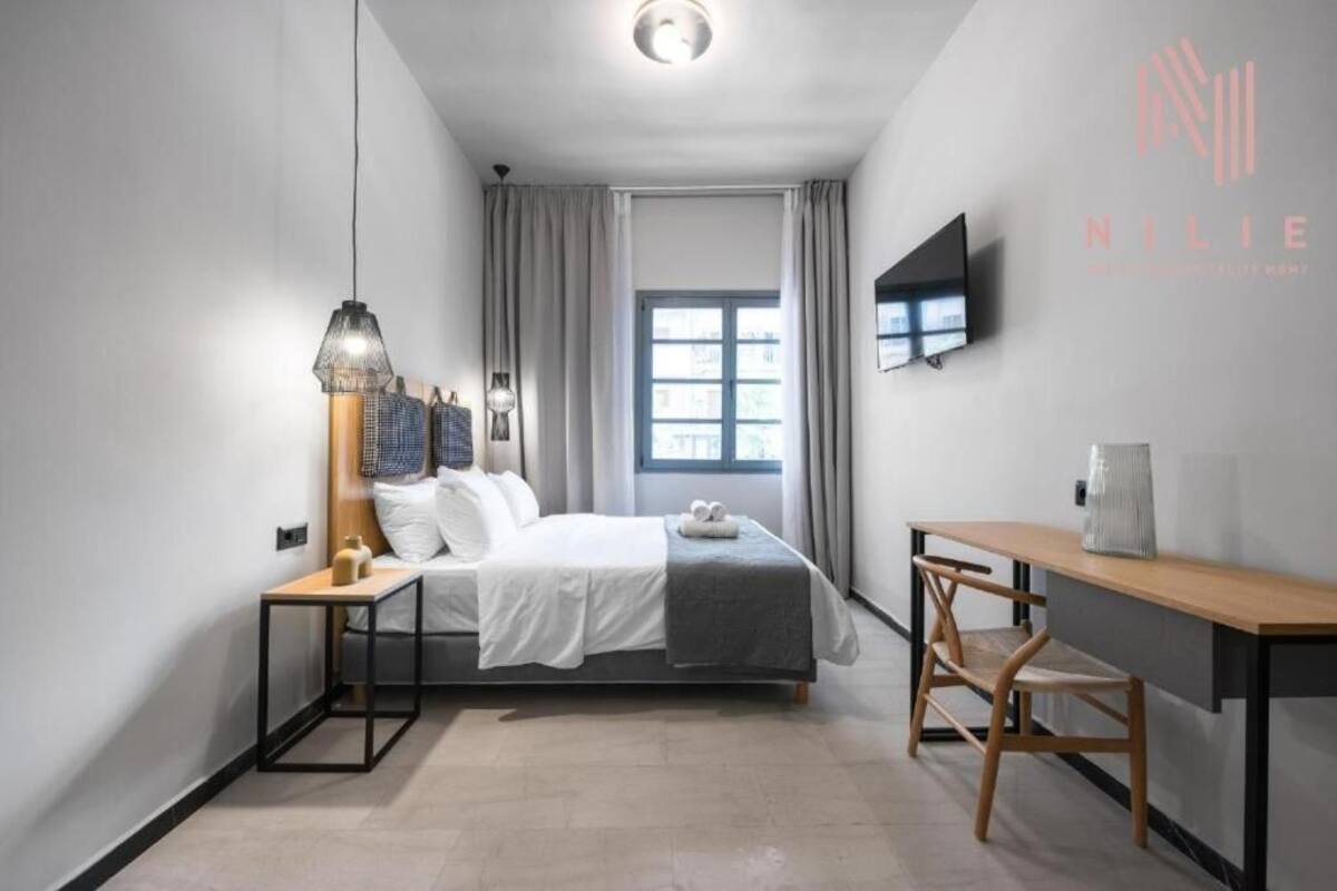 Una habitación de hotel moderna con cama, mesita de noche, lámpara, silla, escritorio y televisor en la pared.