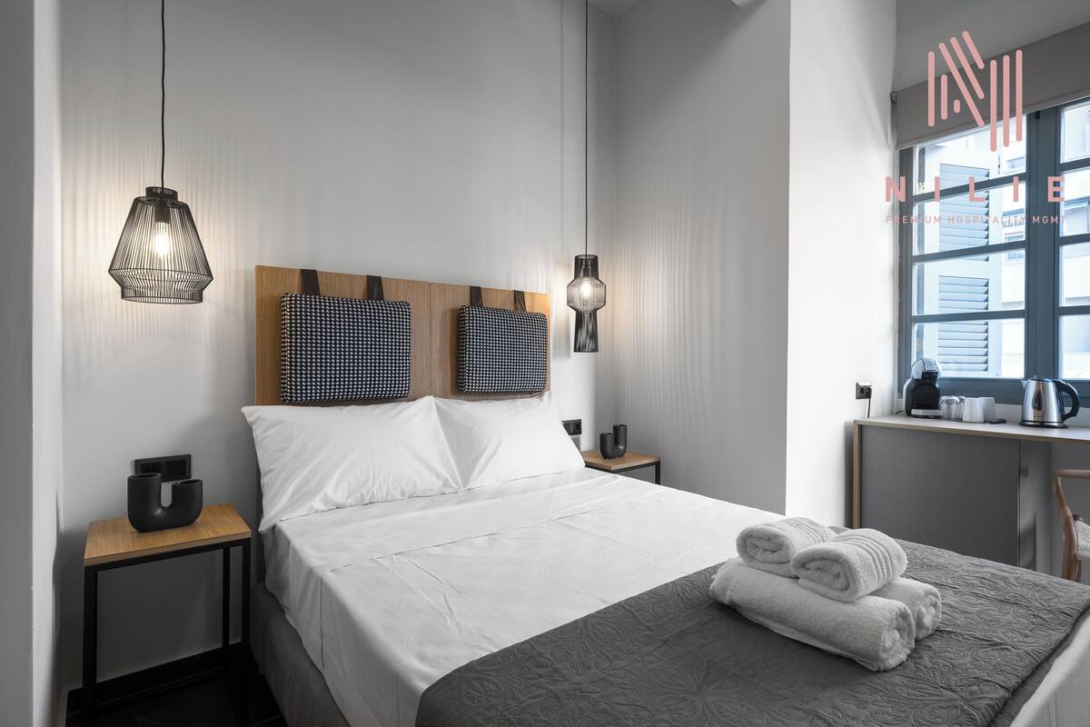Una habitación de hotel moderna con una cama bien hecha, dos mesitas de noche, un escritorio pequeño con una silla y una ventana con persianas.