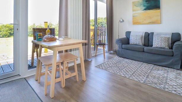 A modern living room with a grey sofa, wooden dining table, and chairs. There is a large window with curtains, a painting on the wall, and a view of greenery outside. - Meadows Edge (Truro)