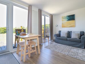 A modern living room with a grey sofa, wooden dining table, and chairs. There is a large window with curtains, a painting on the wall, and a view of greenery outside.