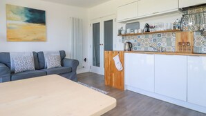 A modern living room with a sofa, a wooden coffee table, and a kitchen area with white cabinets and a tiled backsplash. - Meadows Edge (Truro)