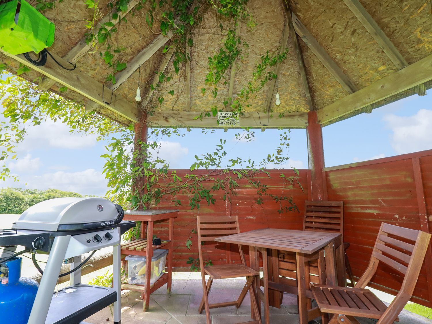 Eine Terrasse im Freien mit Grill, Holztischen und -stühlen sowie einem überdachten Bereich mit Holzboden und hängenden Pflanzen.