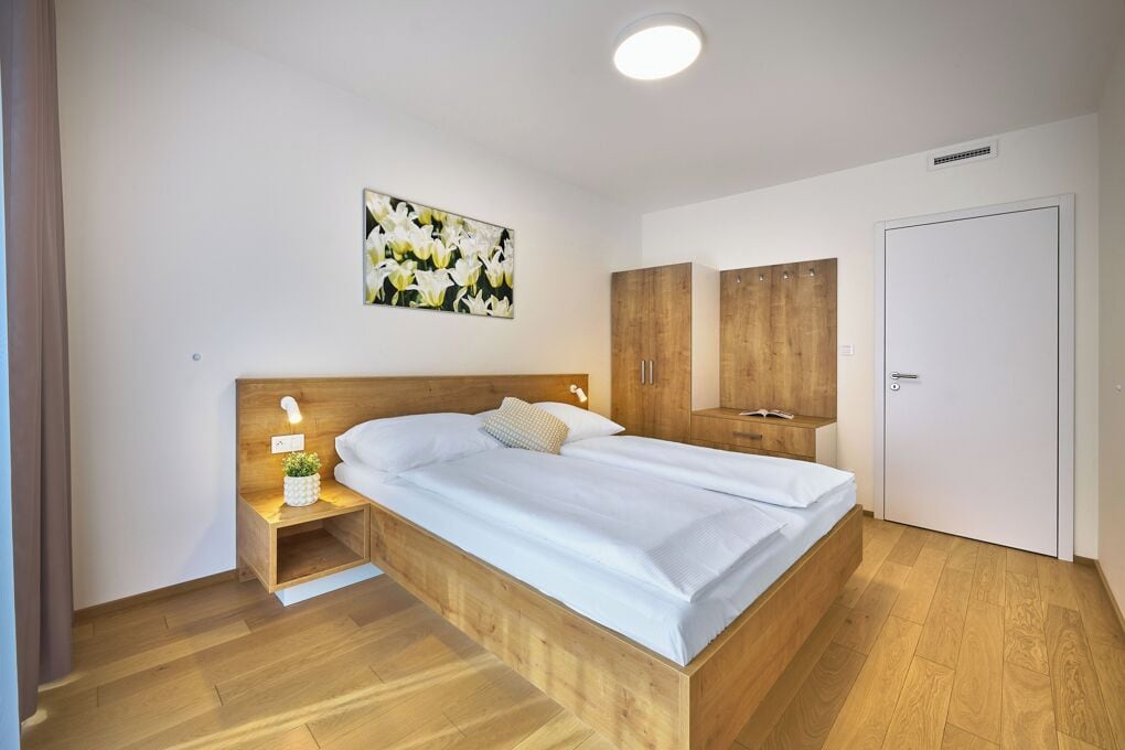 A bedroom with a wooden bed, a nightstand with a lamp, a painting of flowers on the wall, and wooden wardrobes.