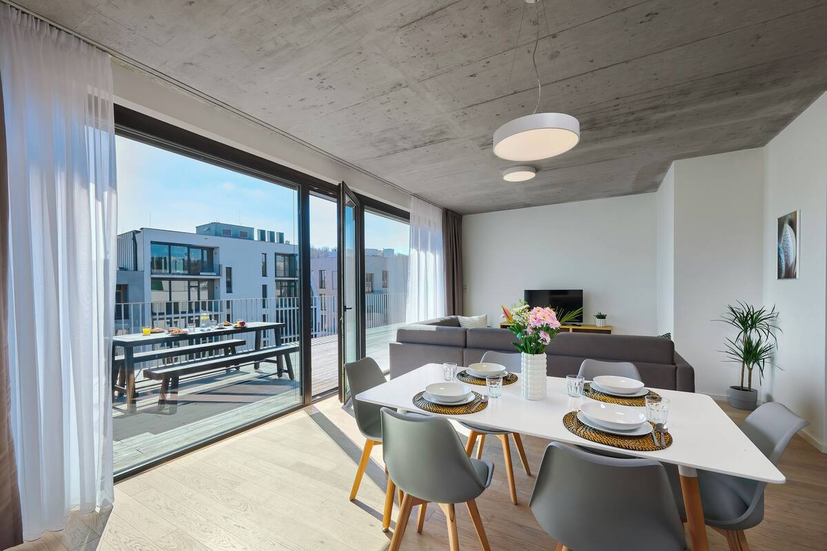 A modern living room with a dining area, large windows, a grey sofa, and a flat-screen TV.