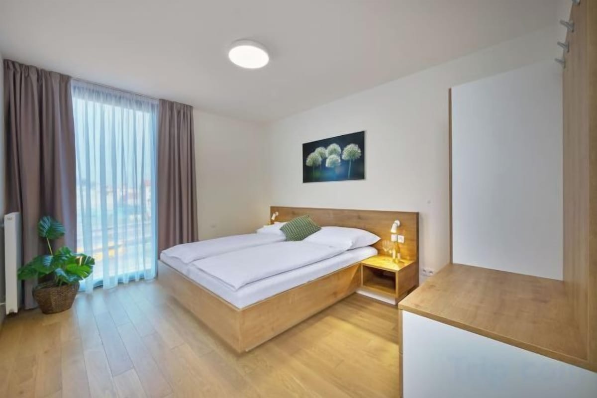 Habitación de hotel con cama de madera, una mesita de noche con lámpara, ventana con cortinas y un cuadro colgado en la pared.