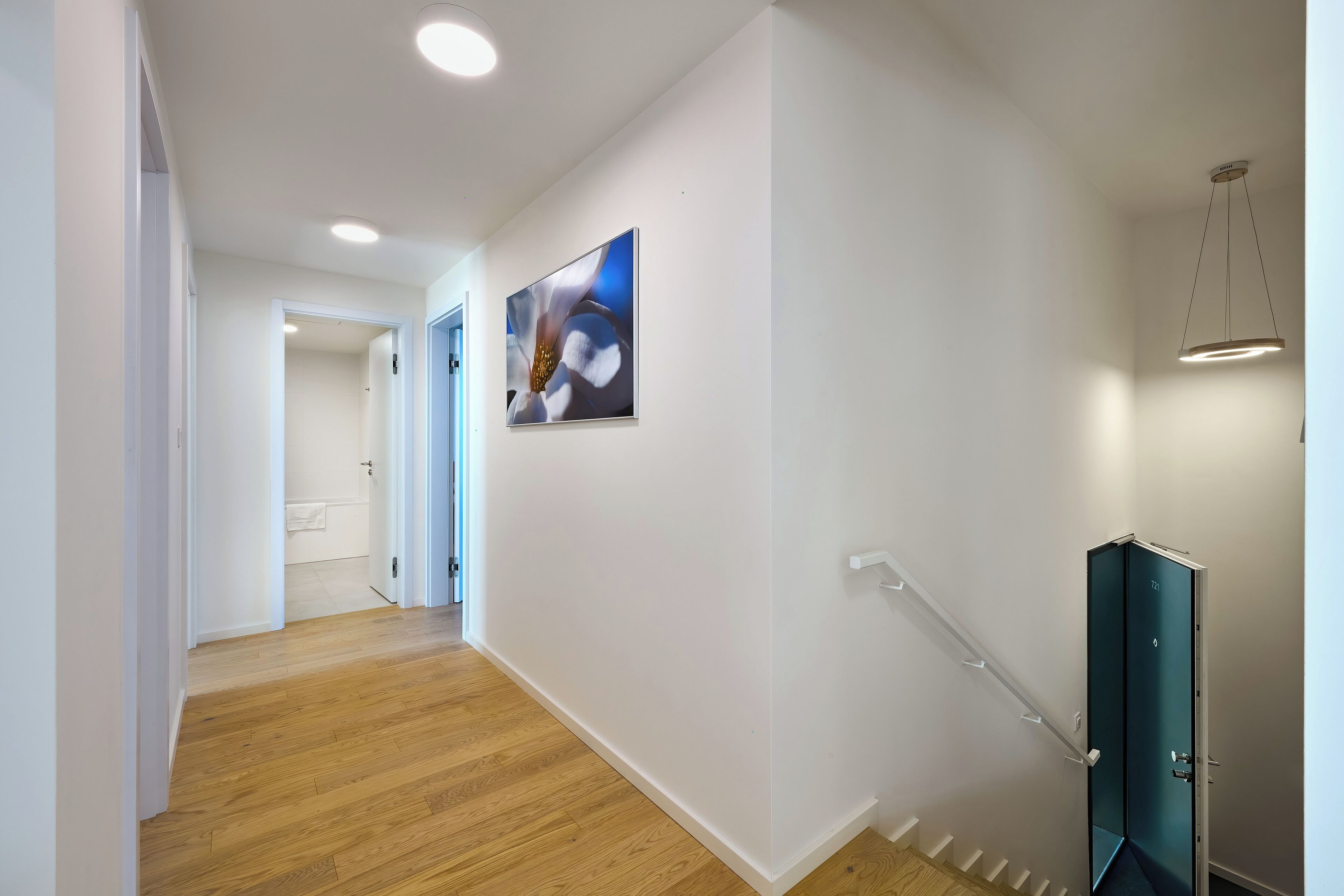 A hallway with wooden flooring, white walls, a ceiling light, a door with a number, and a painting.