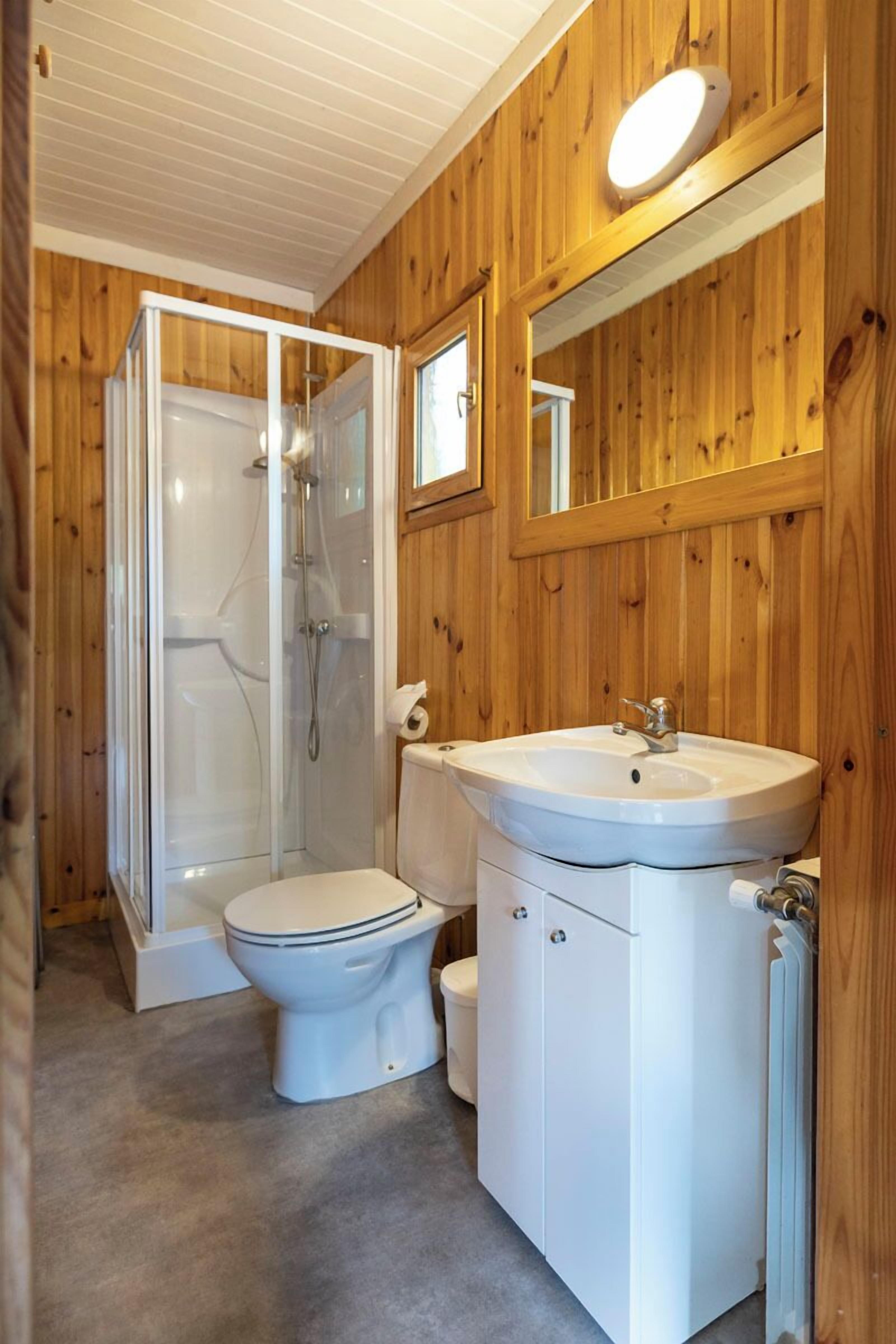 Ein Badezimmer mit Holzwänden, einer Glasdusche, einer weißen Toilette und einem Waschbecken mit Spiegel darüber.