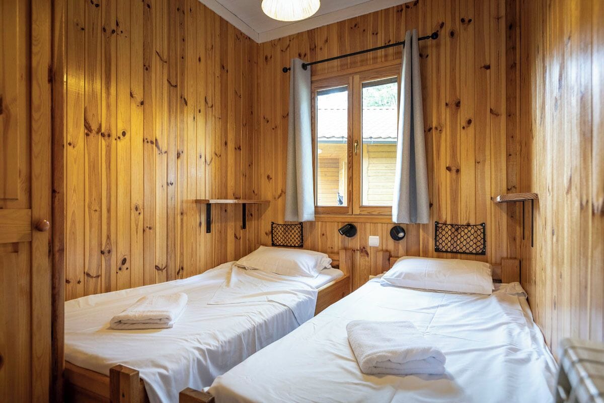Habitació d'una cabana de fusta amb dos llits, una finestra amb cortines i una petita estant.