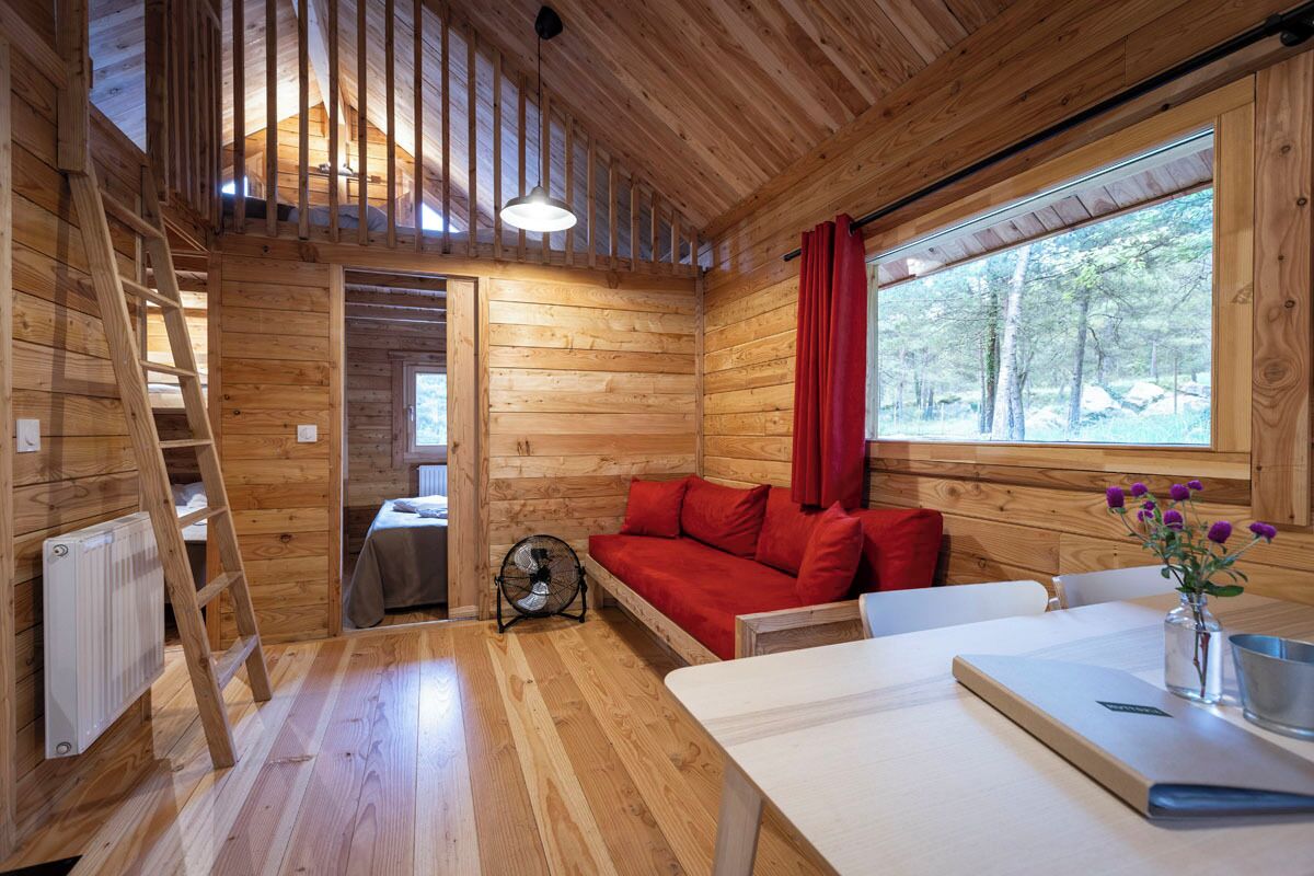 A wooden cabin interior with a loft bed, a sofa, a dining table, and a window with a view of trees.
