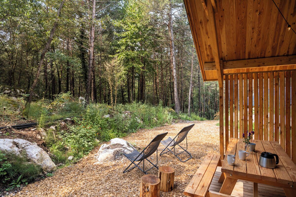 Una terraza de madera con dos sillas plegables y una mesa, rodeada de un bosque.