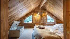 Un dormitorio acogedor en el ático con techo inclinado de madera, una cama con manta color beige, un aparador y dos ventanas que dejan entrar luz natural.