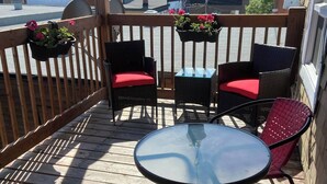 A wooden deck with a round table and two chairs, one with a red cushion. There are hanging flower baskets and a satellite dish.
