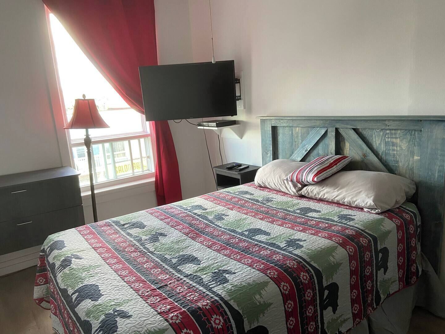 Une chambre à coucher avec un lit, un téléviseur fixé au mur, une table de chevet et une fenêtre avec des rideaux.