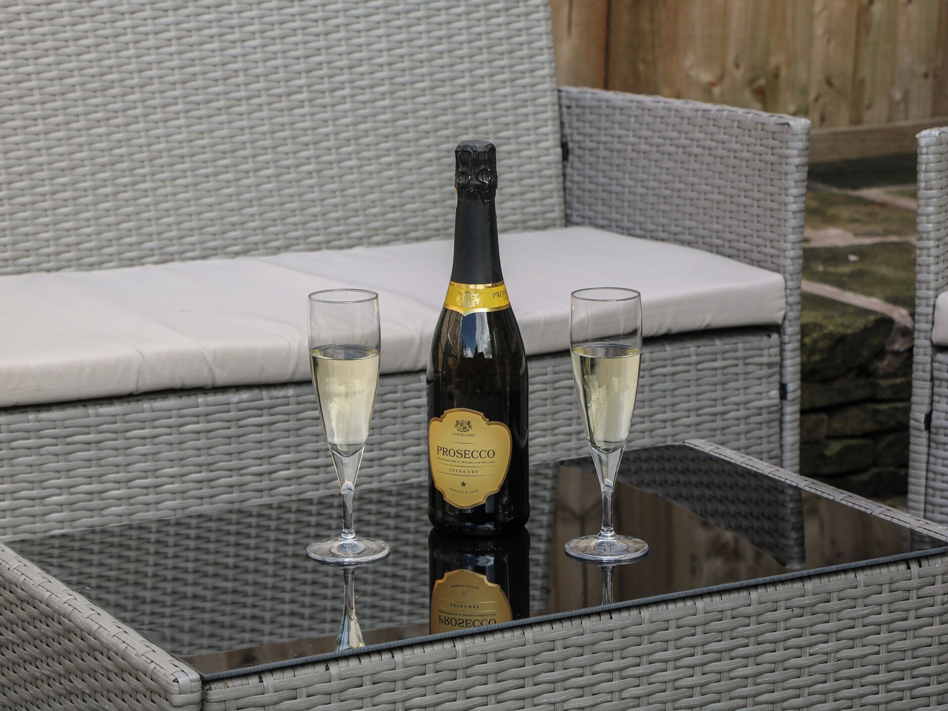 A bottle of Prosecco wine and two glasses on a glass table in an outdoor wicker seating area.