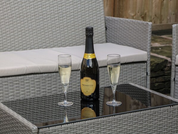 A bottle of Prosecco wine and two glasses on a glass table in an outdoor wicker seating area.