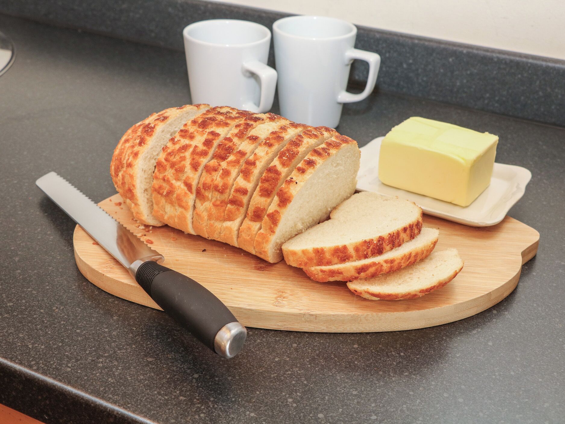 Une miche de pain tranché posée sur une planche à découper en bois, avec un couteau à côté, ainsi qu’une barquette de beurre et deux tasses blanches.