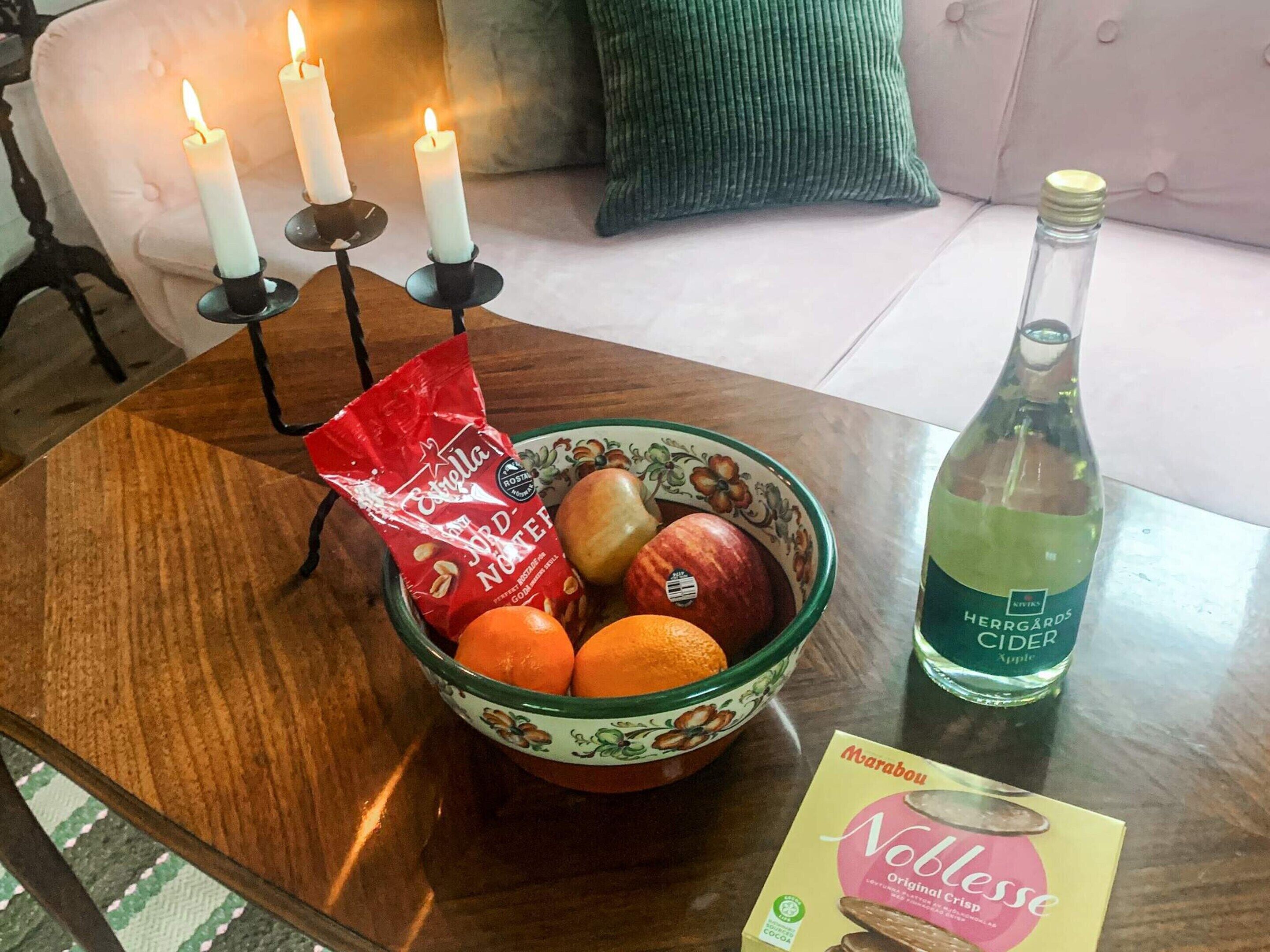 Une table en bois sur laquelle se trouvent un saladier rempli de fruits, une bouteille de cidre, un paquet de chocolat et un chandelier avec des bougies allumées.