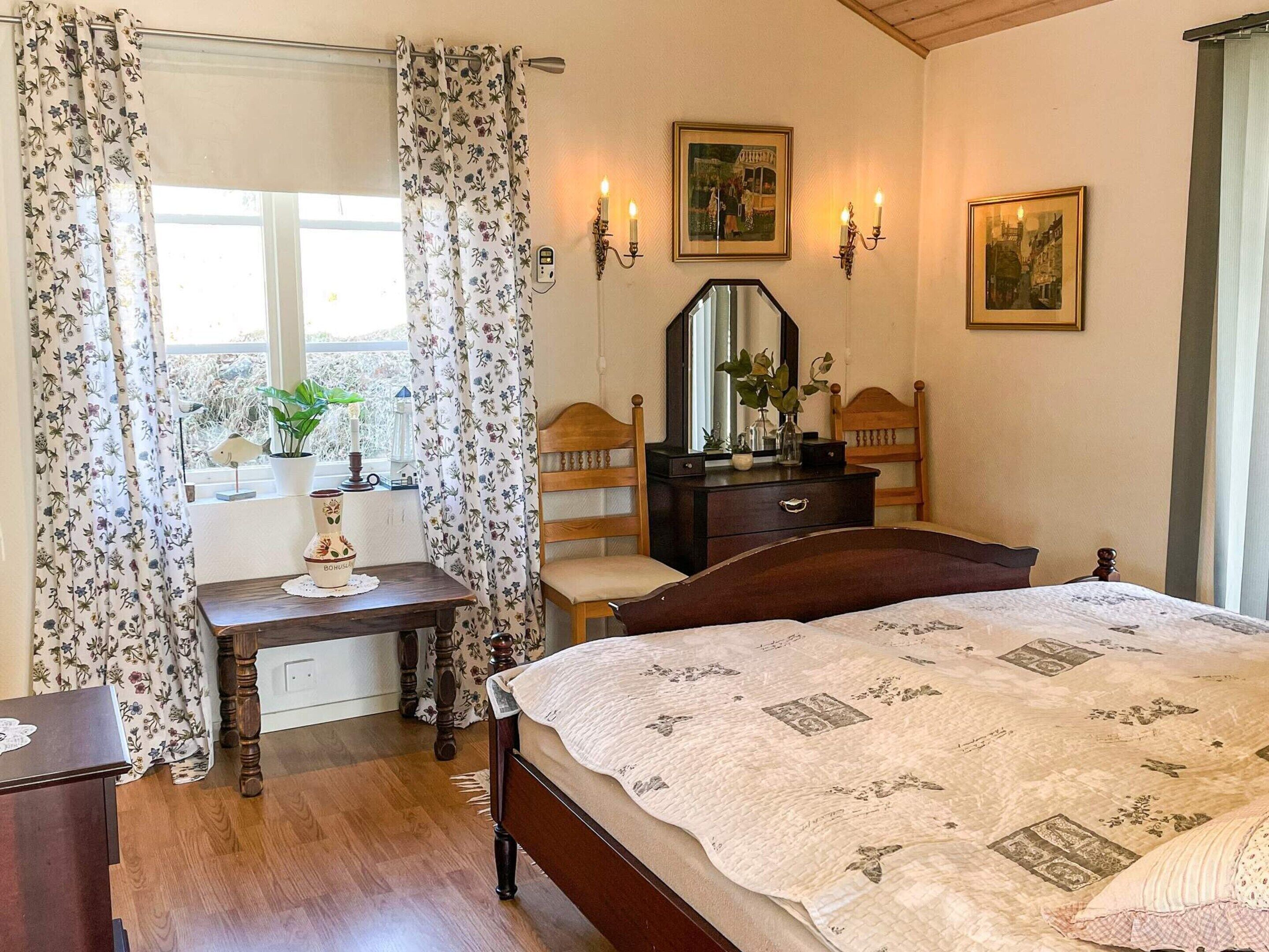 Ein Schlafzimmer mit Bett, Kommode, Spiegel und einem Fenster mit floralem Vorhang.