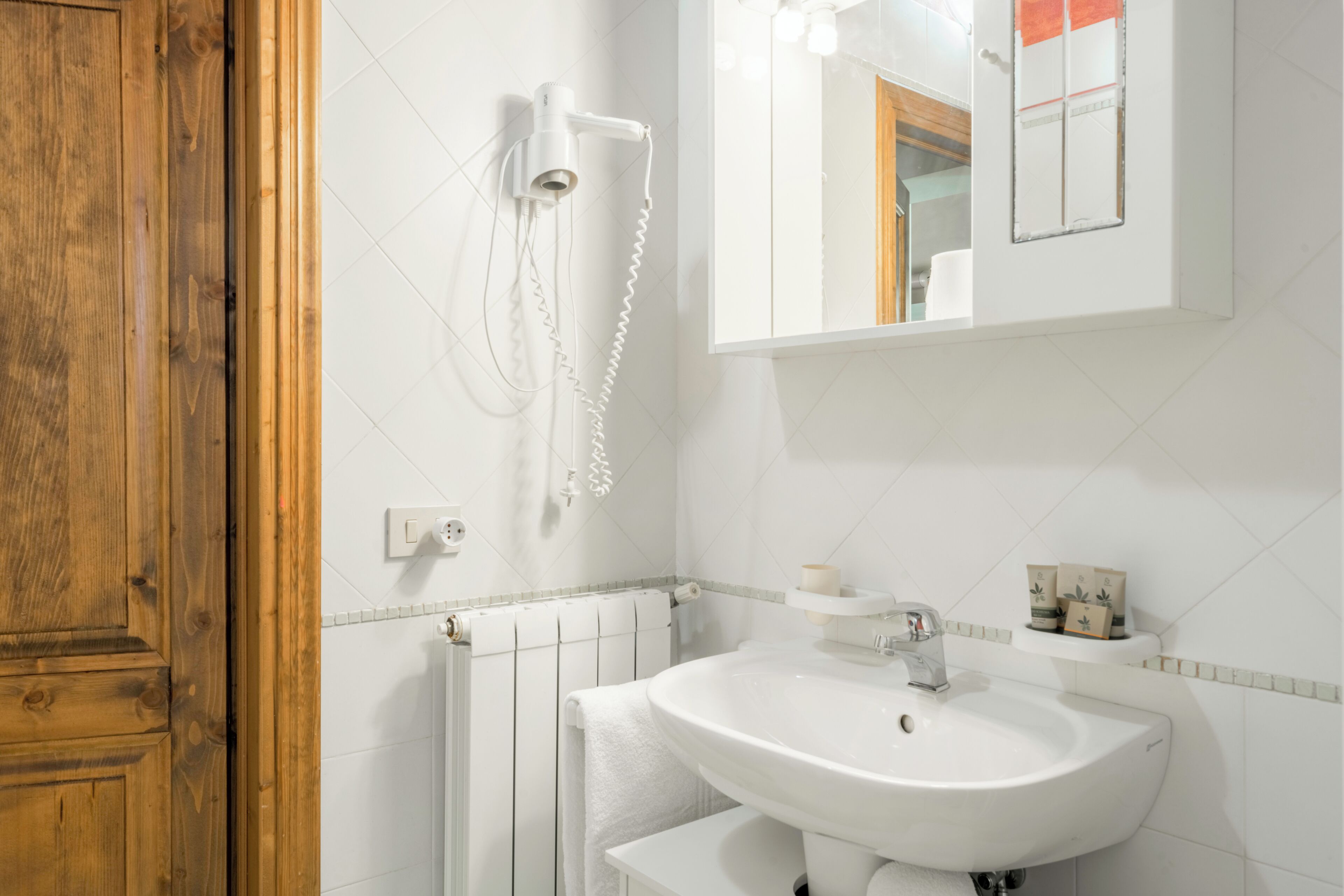 Ein Badezimmer mit Holztür, weißen Fliesen, einem weißen Waschbecken, einem Handtuchhalter, einem Spiegel und einem an der Wand hängenden Handtuch.