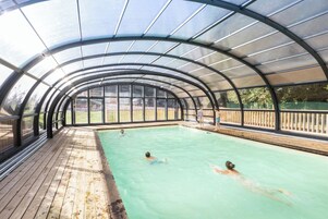 A covered swimming pool with a wooden deck and glass panels.
