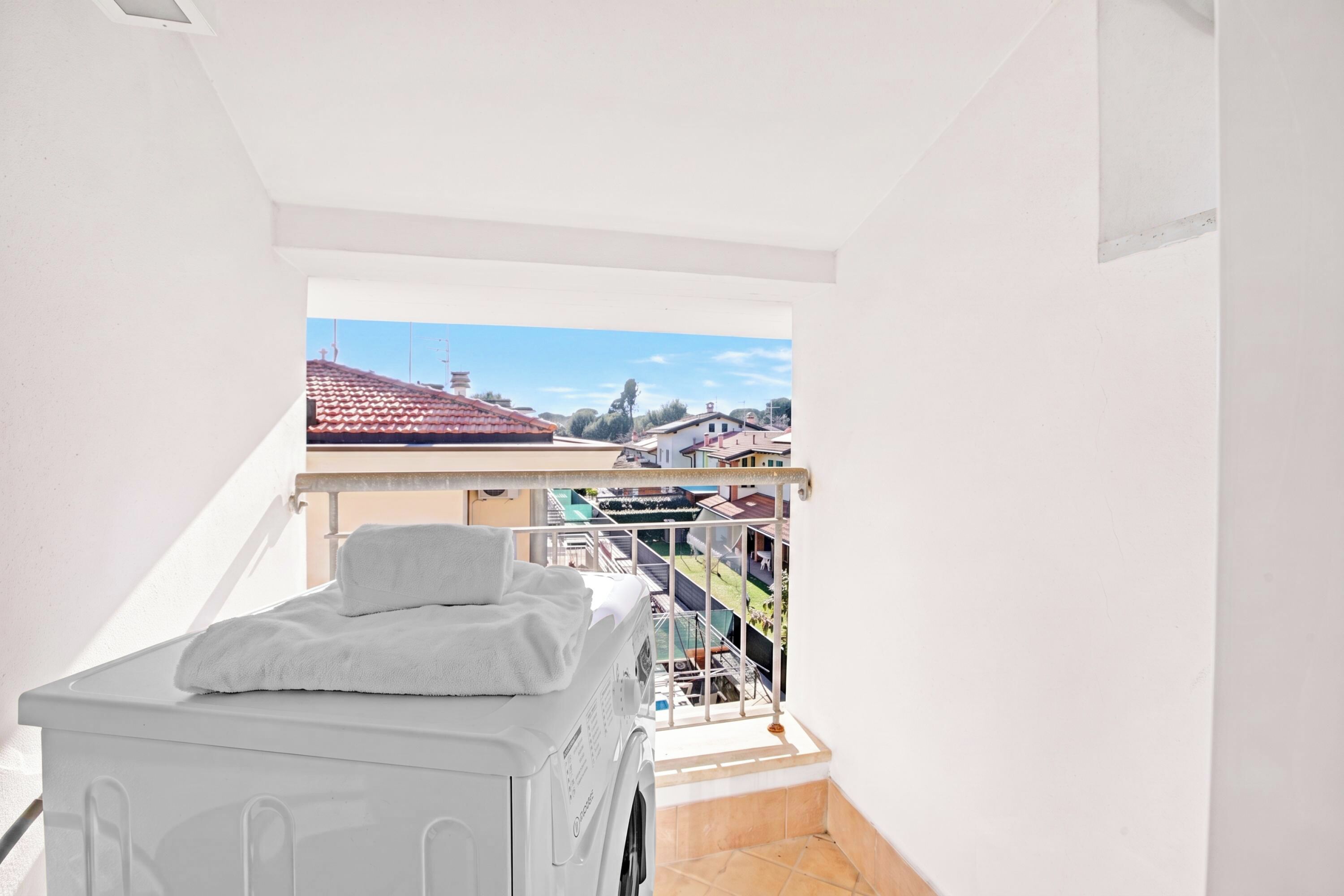 En smal balkon med en hvid vaskemaskine, et håndklæde og udsigt til nabobygninger.