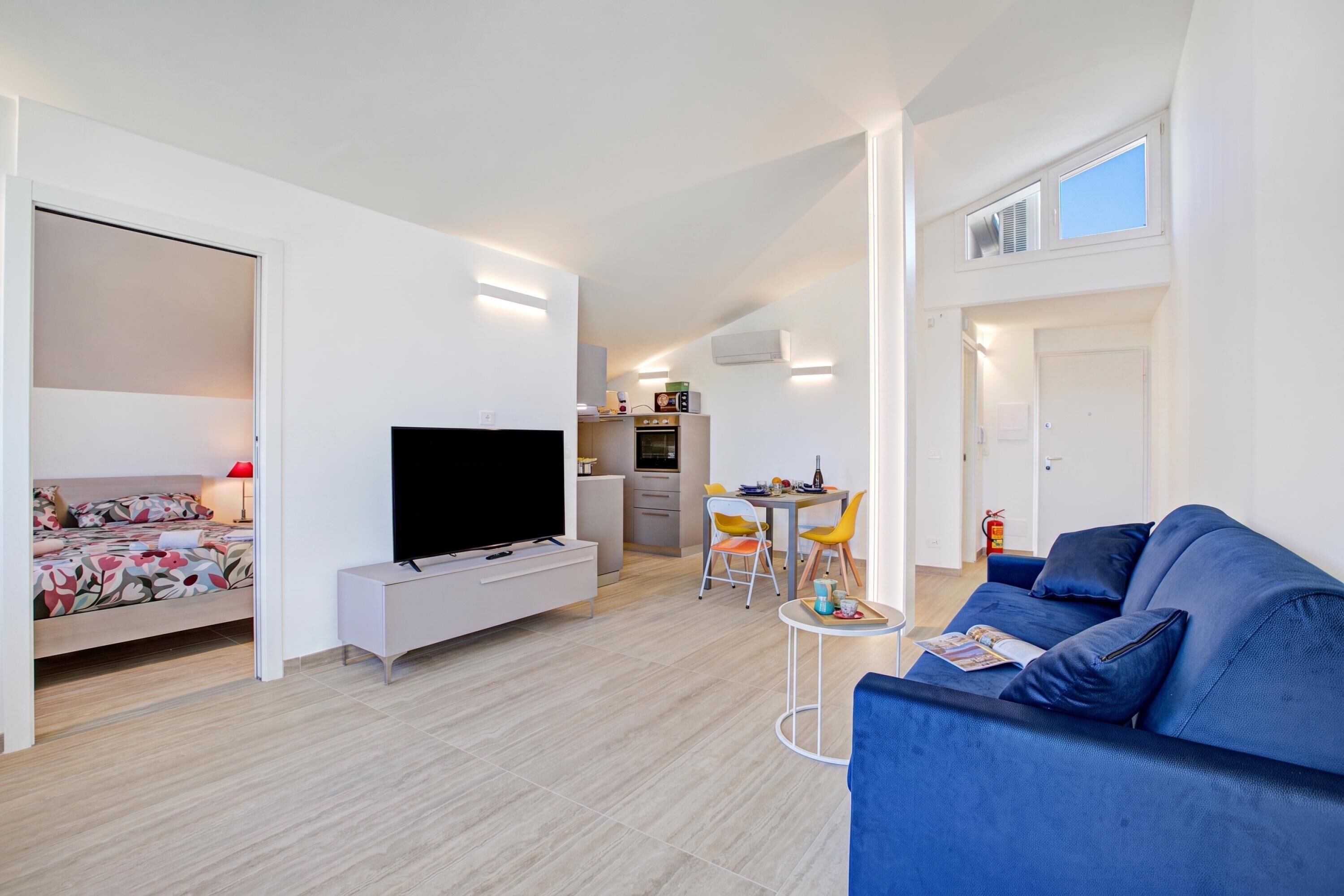 A modern living room with a flat-screen TV, a sofa, a dining area with a table and chairs, and a kitchen in the background.