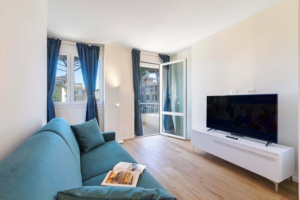 A modern living room with a teal sofa, a flat-screen TV on a white console, and a sliding glass door leading to an outdoor balcony.