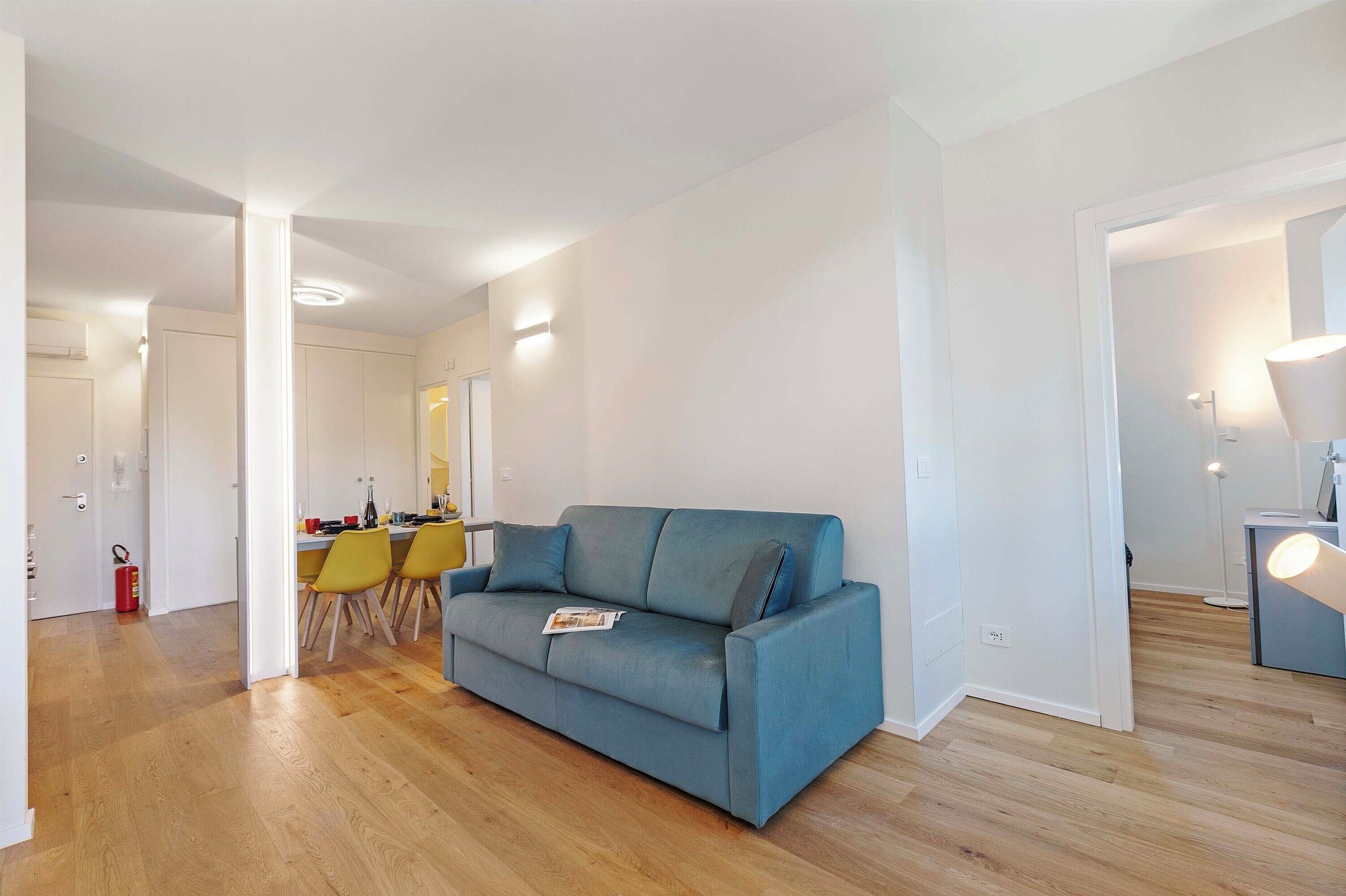 A modern living room with a blue sofa, wooden flooring, and a dining area with yellow chairs.