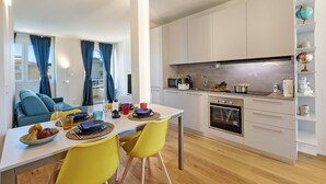 Une cuisine moderne avec des armoires blanches, un four encastré et un four à micro-ondes. On y trouve une table à manger avec des chaises jaunes et, au fond, un canapé bleu.