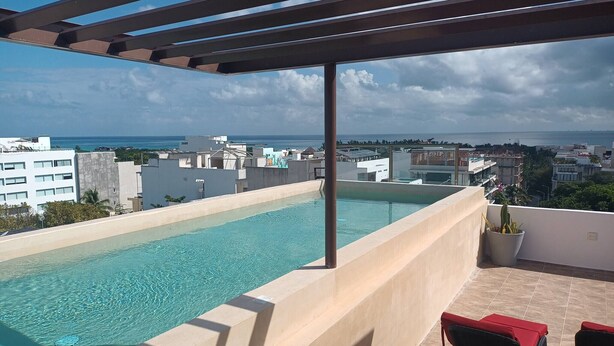 A rooftop pool with clear blue water, surrounded by a pergola and a view of the cityscape and ocean.