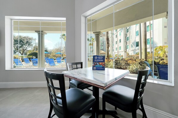 A hotel room with a marble table and four black chairs, large windows with a view of a pool and palm trees, and a sign advertising a 200+ international conference.