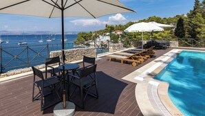 A poolside area with lounge chairs and a table under a large umbrella, overlooking a body of water with boats. - Dream property right on the water (Rogač)