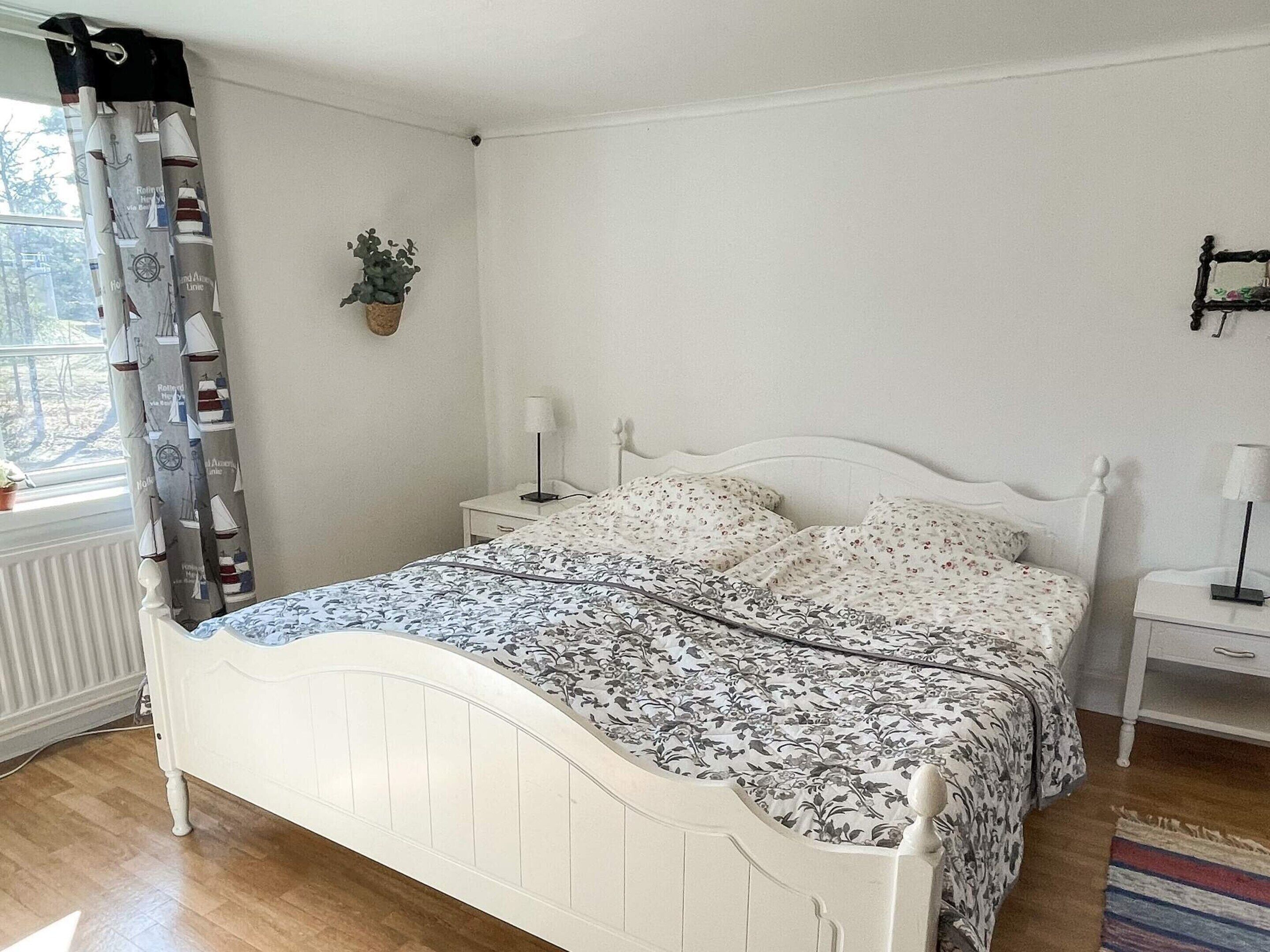 Un lit blanc avec une couvre-lit à motifs floraux, une table de chevet avec une lampe, une fenêtre avec un rideau et une petite plante accrochée au mur.