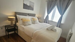 Ein ordentlich bezogenes Bett mit weißer Decke und Kissen, ein Nachttisch mit Lampe, eine Deckenleuchte und ein Fenster mit Vorhängen.