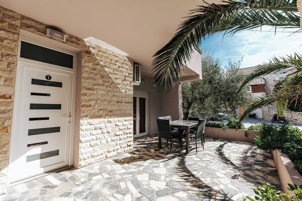 Un bâtiment en pierre avec une porte blanche, une terrasse avec une table et des chaises, et un palmier.