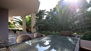 A glass-top table with wicker chairs, a stone-paved patio, and lush greenery in the background. - Apartments Lola (Novalja)