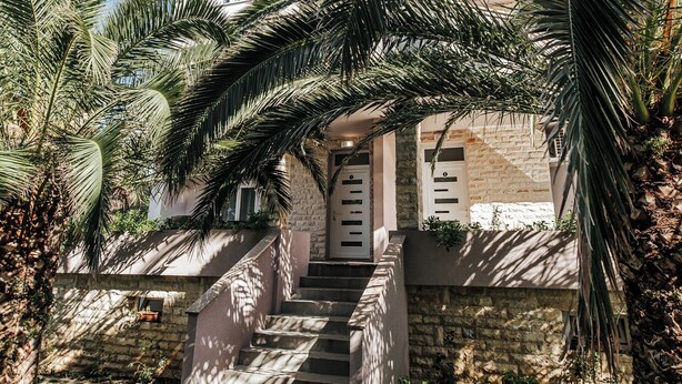 A building with a stone facade, a staircase leading to the entrance, and surrounding palm trees. - Apartments Lola (Novalja)