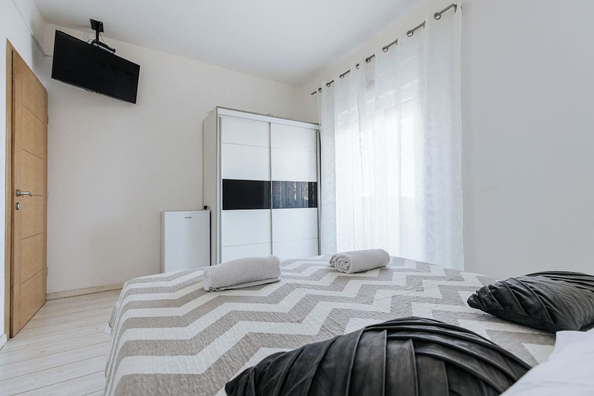 A modern bedroom with a bed, a wardrobe, a TV mounted on the wall, and a window with sheer curtains.