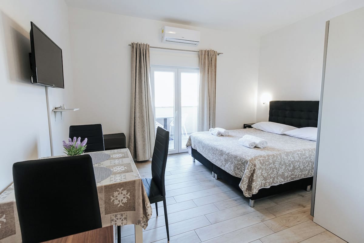 A hotel room with a bed, a TV mounted on the wall, a table with a chair, and a window with curtains.