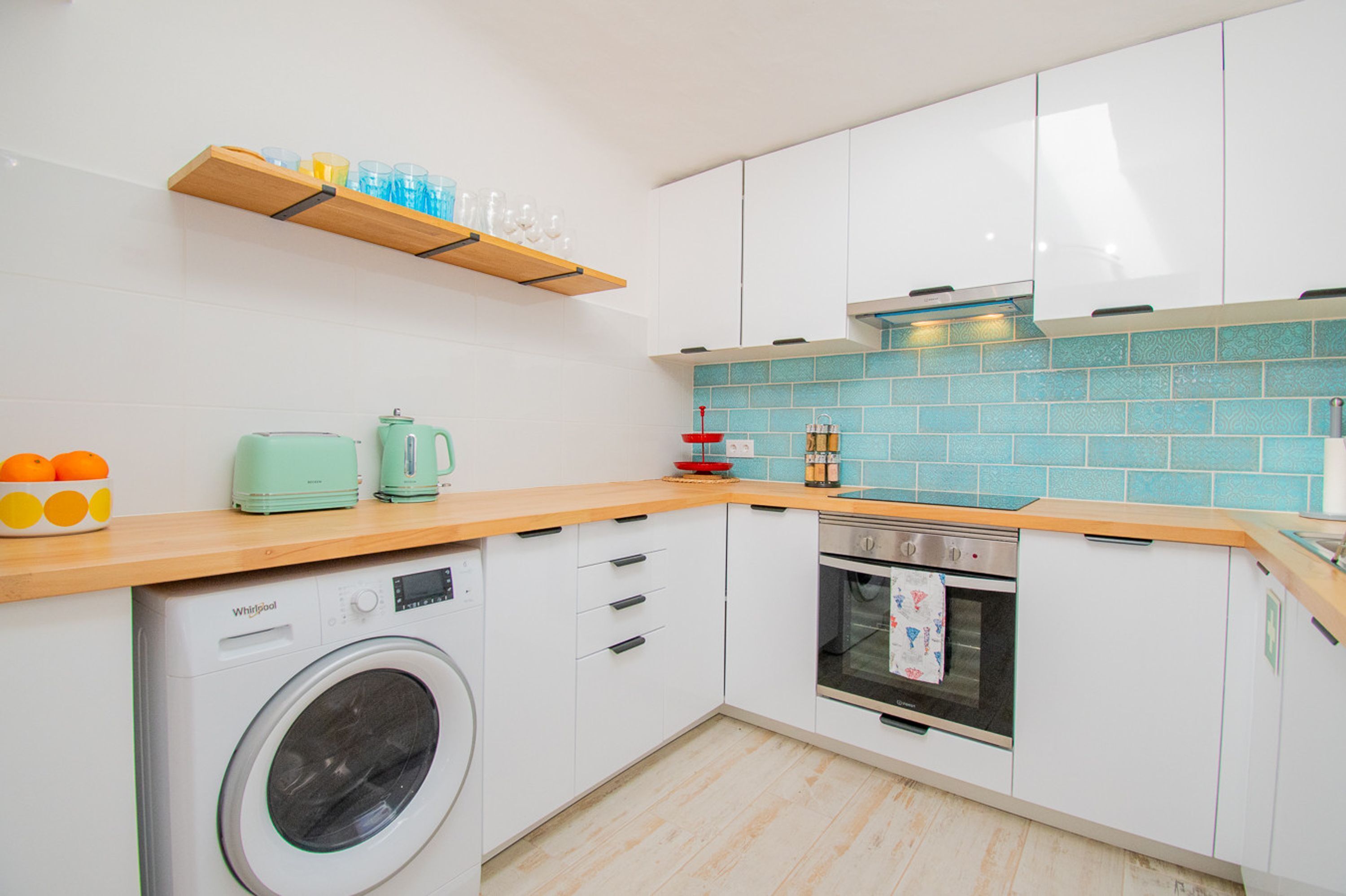 Une cuisine compacte avec des meubles blancs, un lave-linge, une machine à café et un dosseret carrelé.
