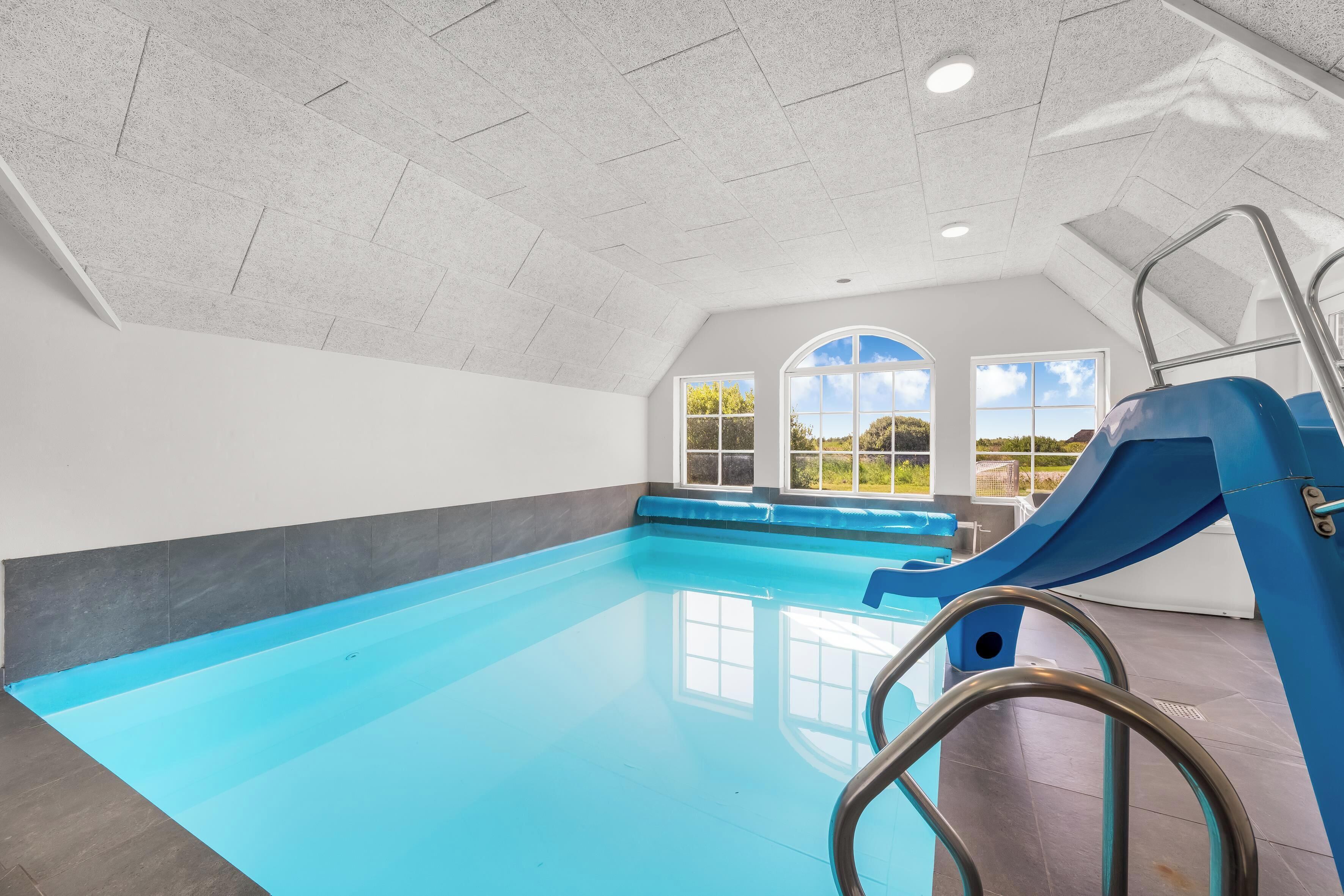 An indoor swimming pool with a slide, large windows, and a view of greenery outside.